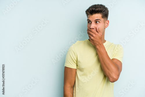 Young caucasian man isolated on blue background thoughtful looking to a copy space covering mouth with hand. © Asier