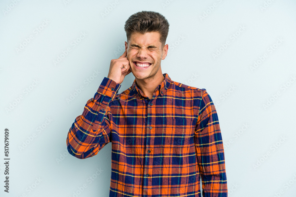 Young caucasian man isolated on blue background covering ears with fingers, stressed and desperate by a loudly ambient. Young caucasian man isolated on blue background covering ears with fingers, stressed and desperate by a loudly ambient.