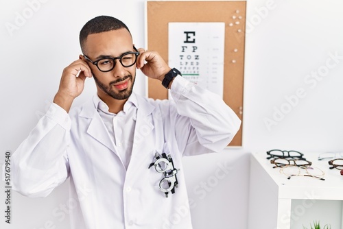 African american optician man standing by eyesight test covering ears with fingers with annoyed expression for the noise of loud music. deaf concept.