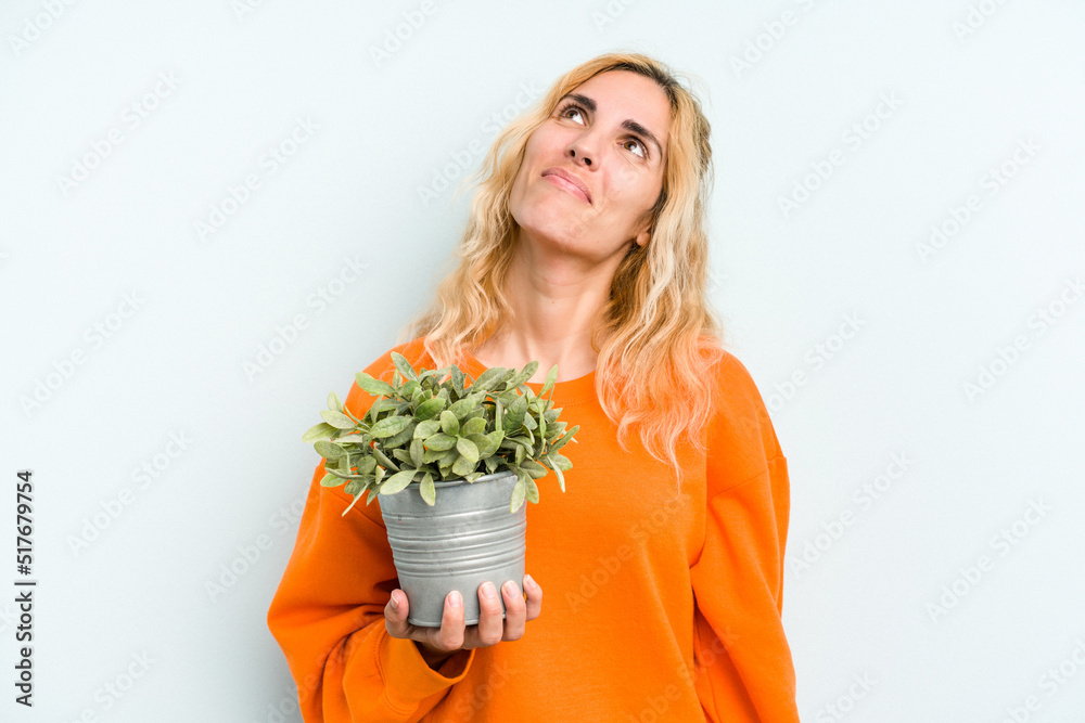 Young caucasian woman holding a plant isolated on blue background dreaming of achieving goals and purposes Young caucasian woman holding a plant isolated on blue background dreaming of achieving goals and purposes