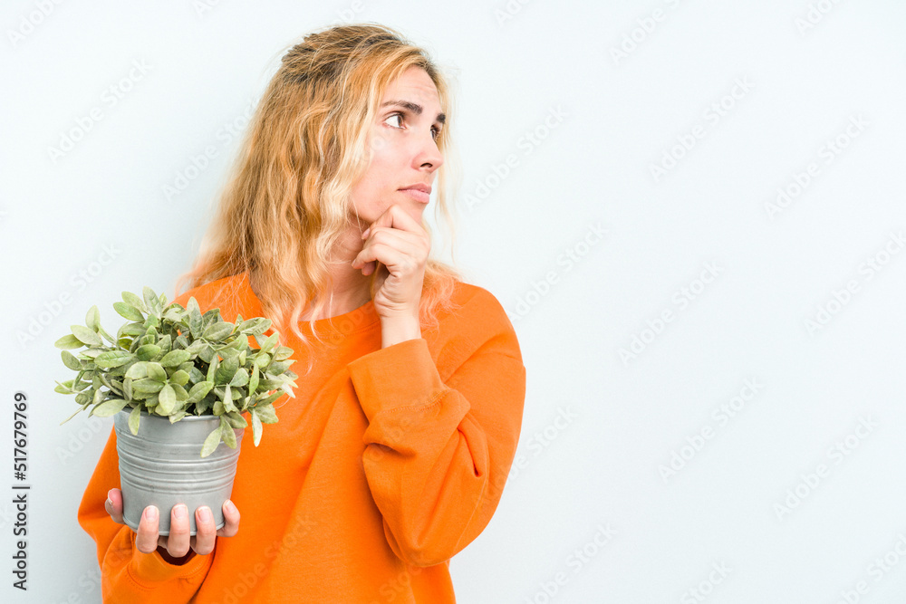 Young caucasian woman holding a plant isolated on blue background looking sideways with doubtful and skeptical expression.