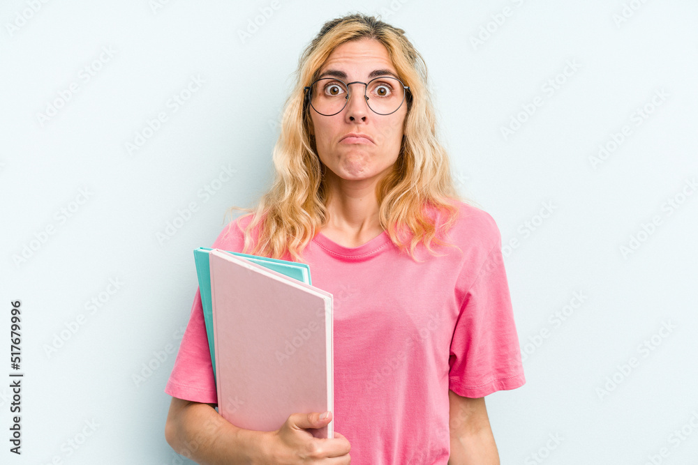 Young student caucasian woman holding books isolated on blue background shrugs shoulders and open eyes confused. Young student caucasian woman holding books isolated on blue background shrugs shoulders and open eyes confused.