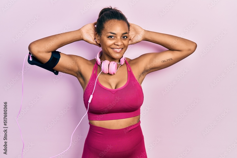 Young african american girl wearing gym clothes and using headphones relaxing and stretching, arms and hands behind head and neck smiling happy