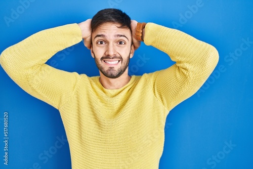 Hispanic man standing over blue background crazy and scared with hands on head, afraid and surprised of shock with open mouth