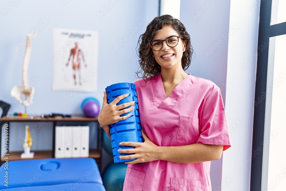 Young latin woman wearing physiotherapist uniform holding foam roller at clinic