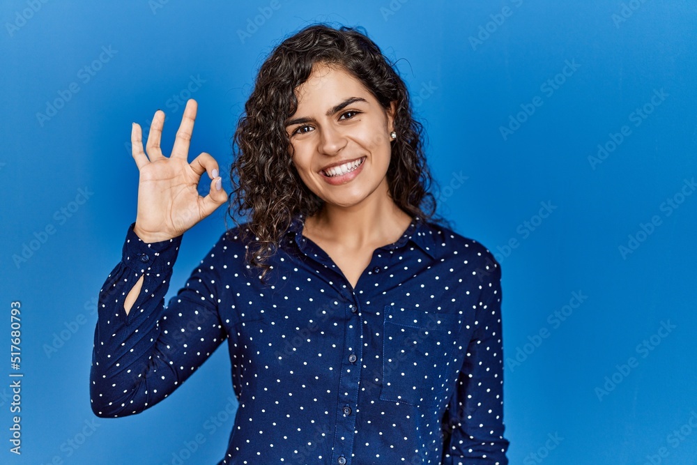 Young brunette woman with curly hair wearing casual clothes over blue background smiling positive doing ok sign with hand and fingers. successful expression. Young brunette woman with curly hair wearing casual clothes over blue background smiling positive doing ok sign with hand and fingers. successful expression.