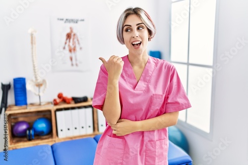Young nurse woman working at pain recovery clinic smiling with happy face looking and pointing to the side with thumb up.