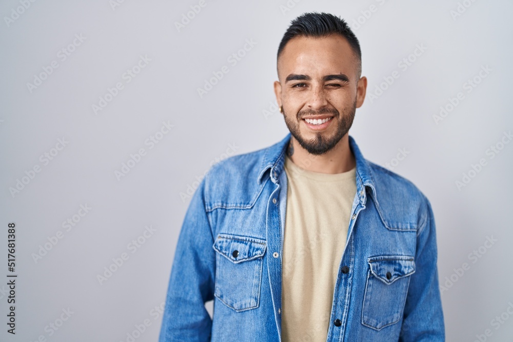 Fototapeta premium Young hispanic man standing over isolated background winking looking at the camera with sexy expression, cheerful and happy face.