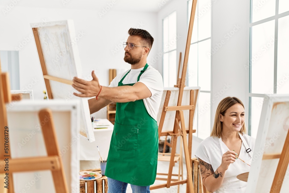 Young caucasian couple smiling happy drawing at art studio. Man standing and holding canvas. Young caucasian couple smiling happy drawing at art studio. Man standing and holding canvas.