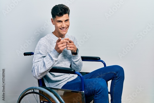 Young hispanic man sitting on wheelchair pointing fingers to camera with happy and funny face. good energy and vibes.