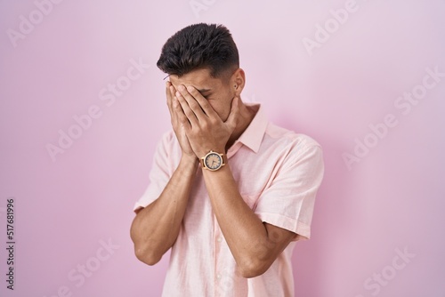 Young hispanic man standing over pink background with sad expression covering face with hands while crying. depression concept.