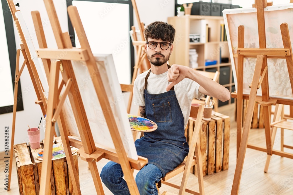 Hispanic man with beard at art studio with angry face, negative sign showing dislike with thumbs down, rejection concept