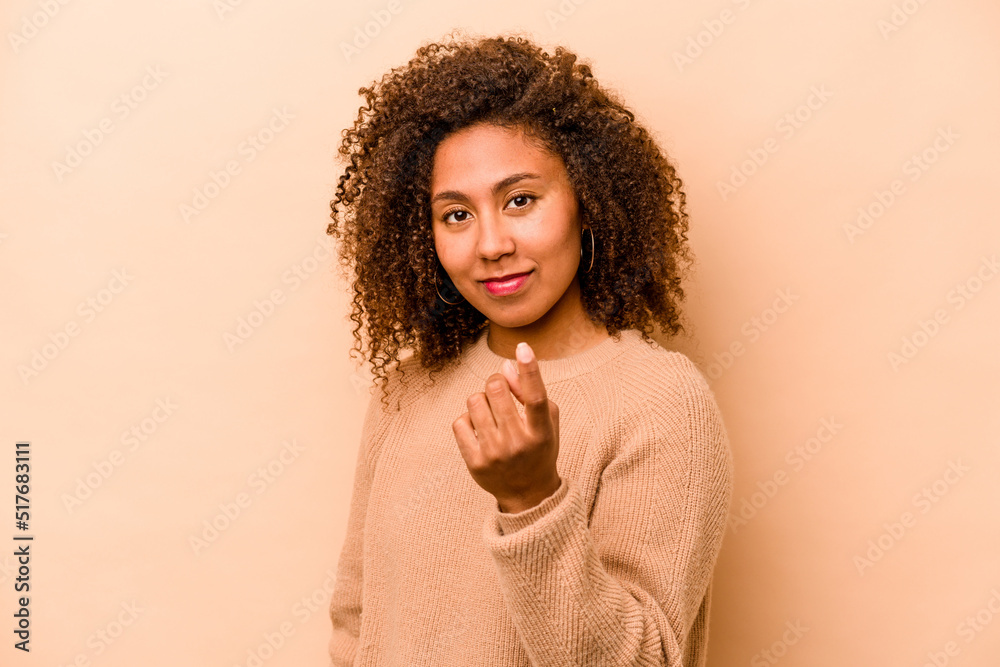 Young African American woman isolated on beige background pointing with finger at you as if inviting come closer. Young African American woman isolated on beige background pointing with finger at you as if inviting come closer.