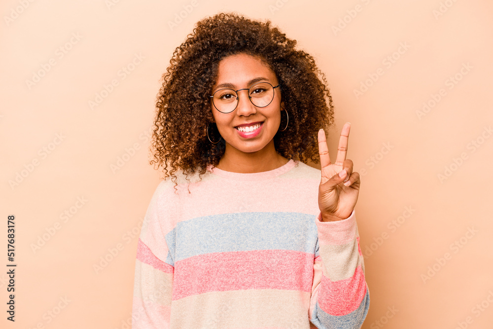 Young African American woman isolated on beige background joyful and carefree showing a peace symbol with fingers. Young African American woman isolated on beige background joyful and carefree showing a peace symbol with fingers.