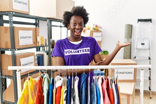 African young woman wearing volunteer t shirt at donations stand amazed and smiling to the camera while presenting with hand and pointing with finger.