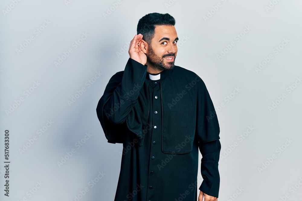 Handsome hispanic man with beard wearing catholic priest robe smiling with hand over ear listening an hearing to rumor or gossip. deafness concept. Handsome hispanic man with beard wearing catholic priest robe smiling with hand over ear listening an hearing to rumor or gossip. deafness concept.