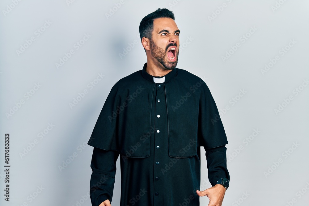 Obraz premium Handsome hispanic man with beard wearing catholic priest robe angry and mad screaming frustrated and furious, shouting with anger. rage and aggressive concept.