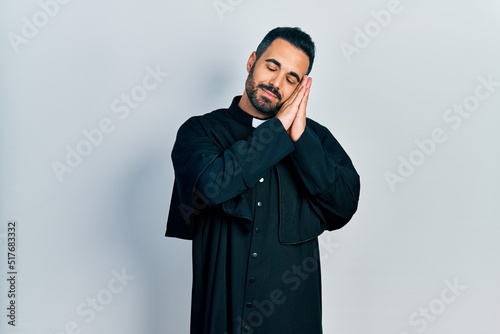 Handsome hispanic man with beard wearing catholic priest robe sleeping tired dreaming and posing with hands together while smiling with closed eyes.