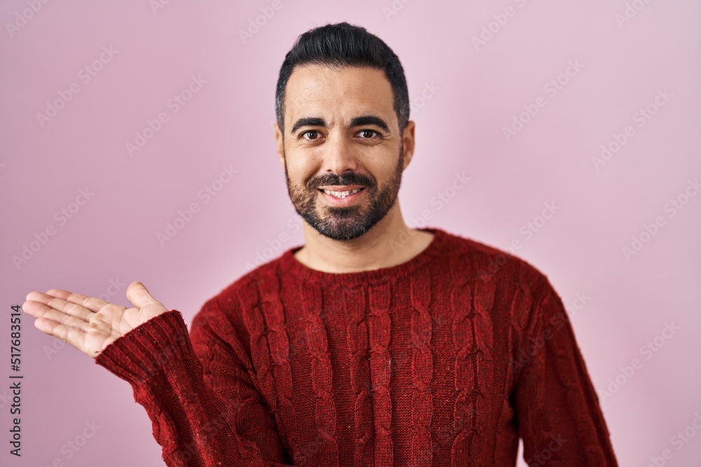 Fototapeta premium Young hispanic man with beard wearing casual sweater over pink background smiling cheerful presenting and pointing with palm of hand looking at the camera.