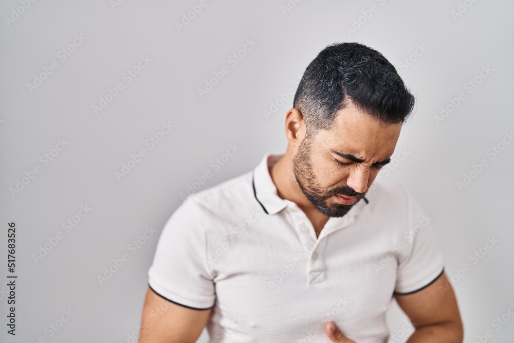 Young hispanic man with beard wearing casual clothes over white background with hand on stomach because indigestion, painful illness feeling unwell. ache concept. Young hispanic man with beard wearing casual clothes over white background with hand on stomach because indigestion, painful illness feeling unwell. ache concept.