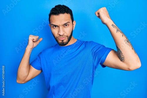 Hispanic man with beard wearing casual blue t shirt showing arms muscles smiling proud. fitness concept.
