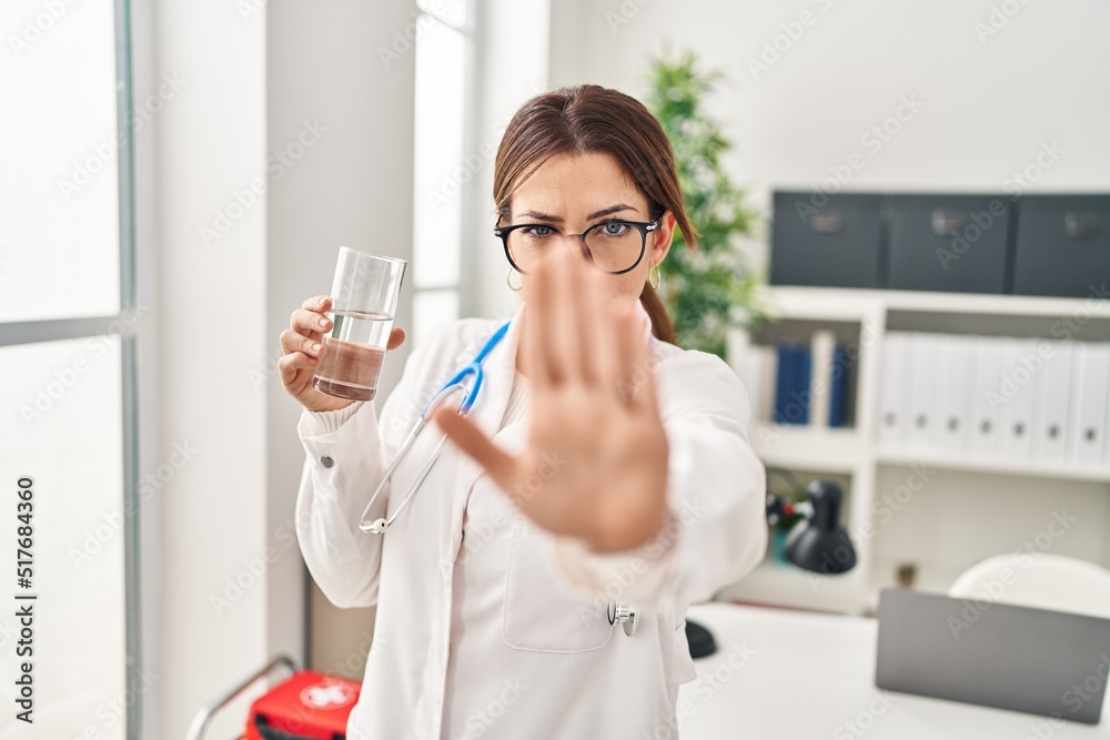 Young brunette doctor woman holding glass of water with open hand doing stop sign with serious and confident expression, defense gesture Young brunette doctor woman holding glass of water with open hand doing stop sign with serious and confident expression, defense gesture