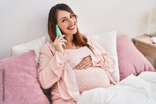 Young pregnant woman talking on the smartphone sitting on bed at bedroom