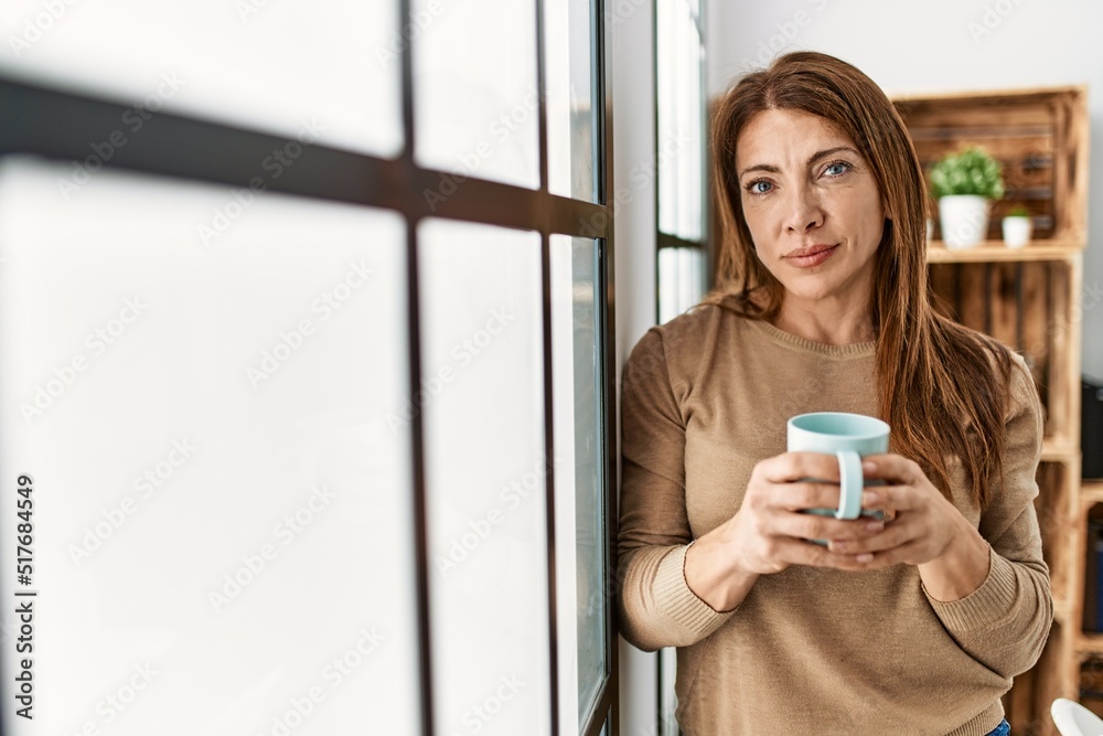 Middle age caucasian woman with serious expression drinking coffee standing at home. Middle age caucasian woman with serious expression drinking coffee standing at home.