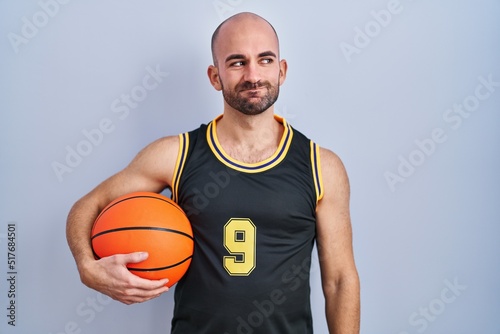 Young bald man with beard wearing basketball uniform holding ball smiling looking to the side and staring away thinking.