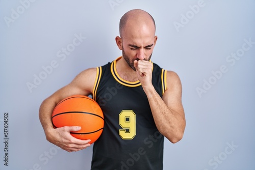 Young bald man with beard wearing basketball uniform holding ball feeling unwell and coughing as symptom for cold or bronchitis. health care concept.
