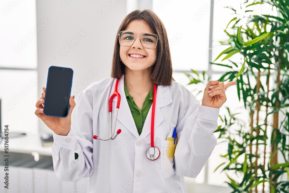 Young hispanic doctor woman showing smartphone screen smiling happy pointing with hand and finger to the side Young hispanic doctor woman showing smartphone screen smiling happy pointing with hand and finger to the side