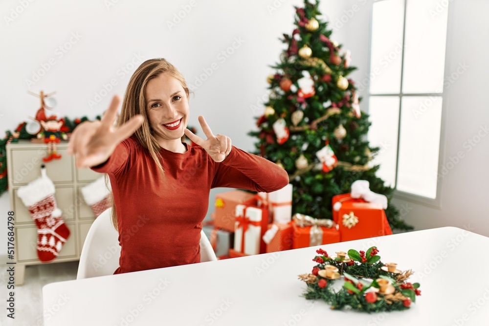 Caucasian young blonde woman sitting on the table by christmas tree smiling looking to the camera showing fingers doing victory sign. number two. Caucasian young blonde woman sitting on the table by christmas tree smiling looking to the camera showing fingers doing victory sign. number two.
