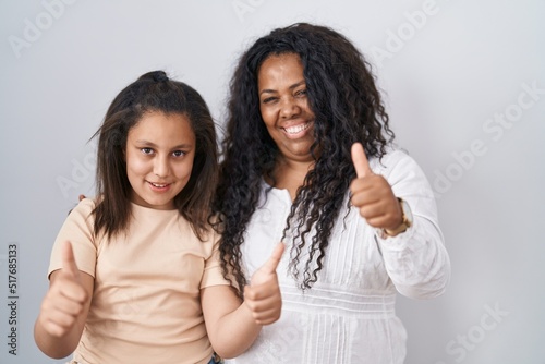 Mother and young daughter standing over white background approving doing positive gesture with hand  thumbs up smiling and happy for success. winner gesture.