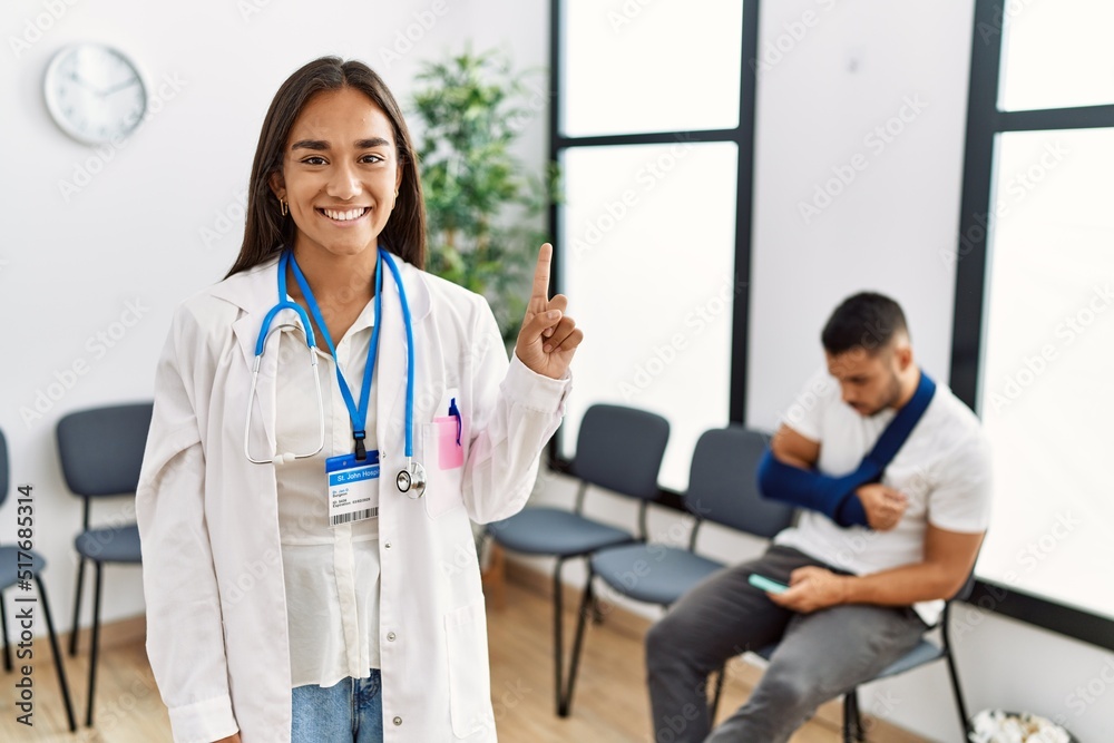 Fototapeta premium Young asian doctor woman at waiting room with a man with a broken arm showing and pointing up with finger number one while smiling confident and happy.