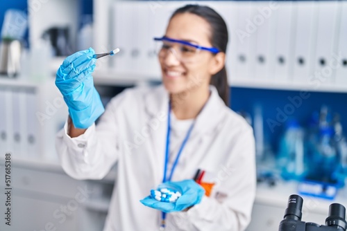 Young hispanic woman scientist holding pills with tweezer at laboratory