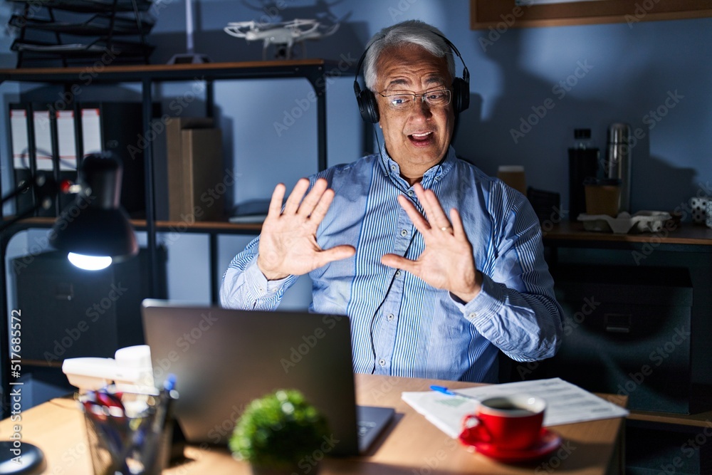 Fototapeta premium Hispanic senior man wearing call center agent headset at night afraid and terrified with fear expression stop gesture with hands, shouting in shock. panic concept.