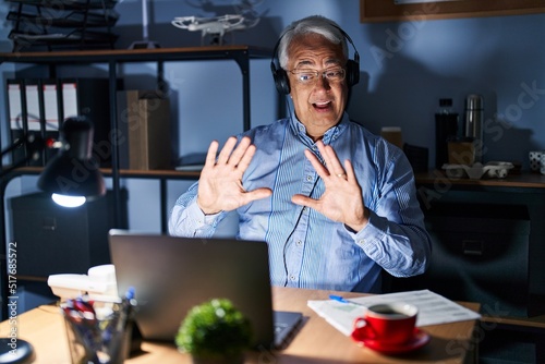 Hispanic senior man wearing call center agent headset at night afraid and terrified with fear expression stop gesture with hands, shouting in shock. panic concept.