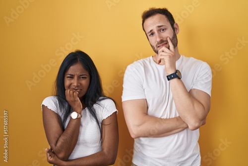 Interracial couple standing over yellow background looking stressed and nervous with hands on mouth biting nails. anxiety problem.