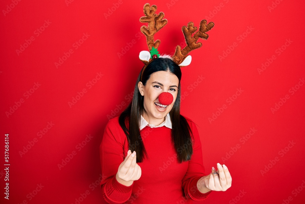 Young hispanic woman wearing deer christmas hat and red nose doing money gesture with hands, asking for salary payment, millionaire business