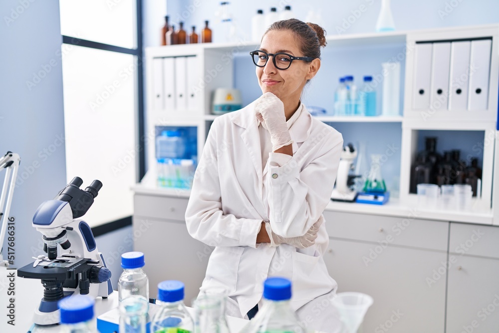 Young woman working at scientist laboratory smiling looking confident at the camera with crossed arms and hand on chin. thinking positive.