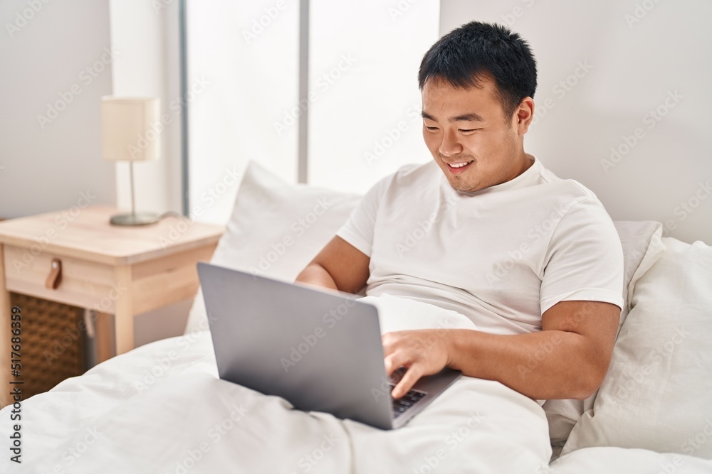 Young chinese man using laptop sitting on bed at bedroom Young chinese man using laptop sitting on bed at bedroom