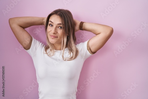 Blonde caucasian woman standing over pink background relaxing and stretching  arms and hands behind head and neck smiling happy