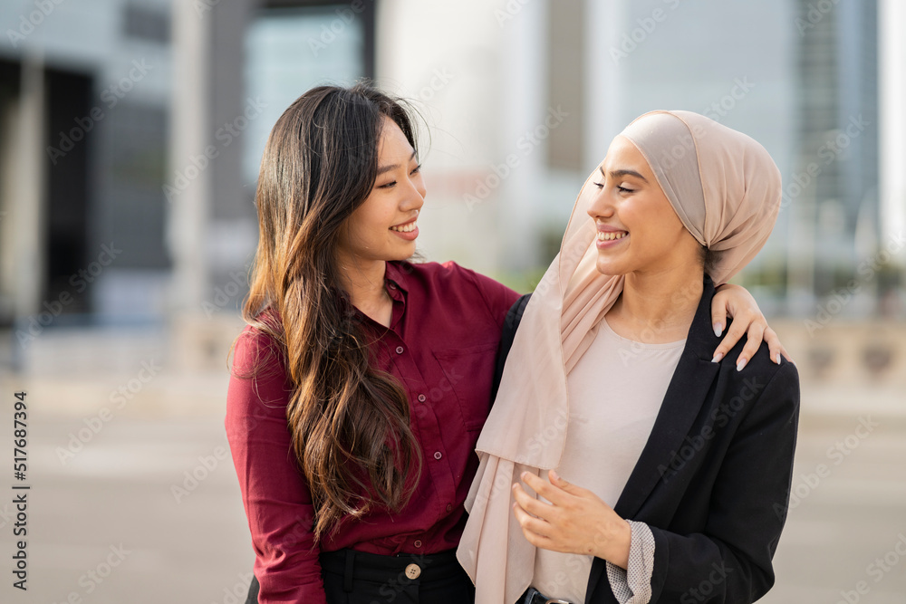 multiracial businesswomen friends hug outside, asian woman with muslim woman in buildings. multiracial businesswomen friends hug outside, asian woman with muslim woman in buildings.