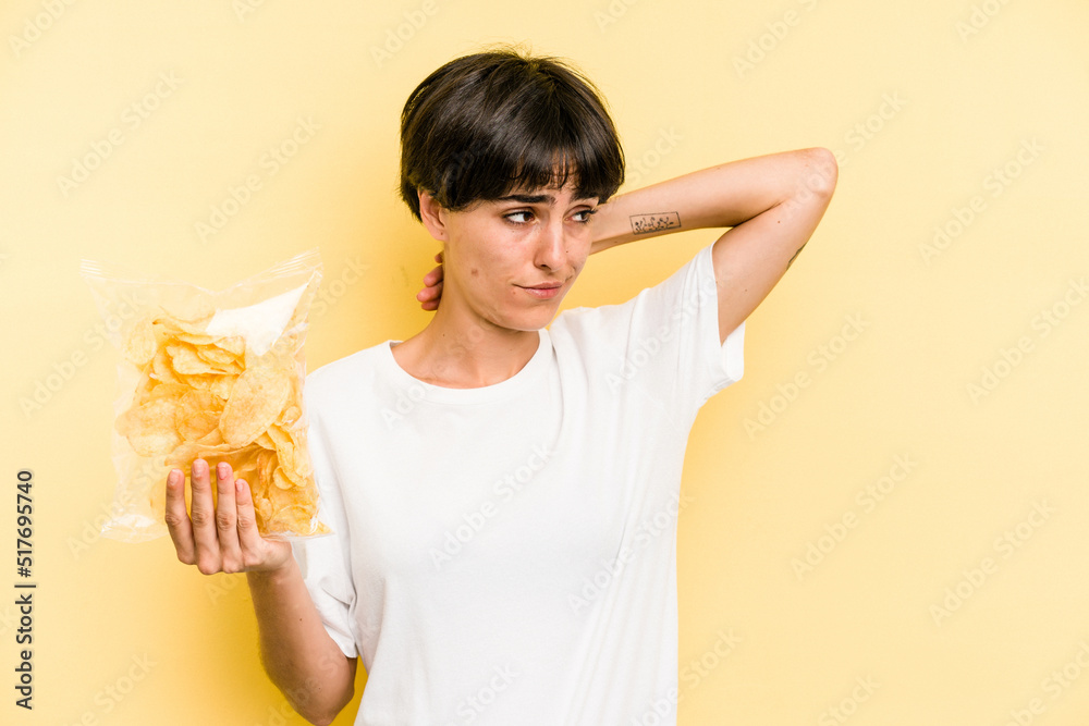 Young caucasian man holding crisps isolated on yellow background touching back of head, thinking and making a choice. Young caucasian man holding crisps isolated on yellow background touching back of head, thinking and making a choice.