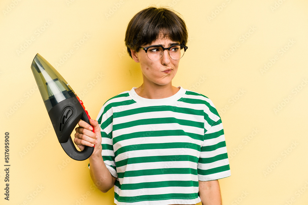 Young caucasian woman holding a hand vacuum cleaner isolated on yellow background confused, feels doubtful and unsure. Young caucasian woman holding a hand vacuum cleaner isolated on yellow background confused, feels doubtful and unsure.