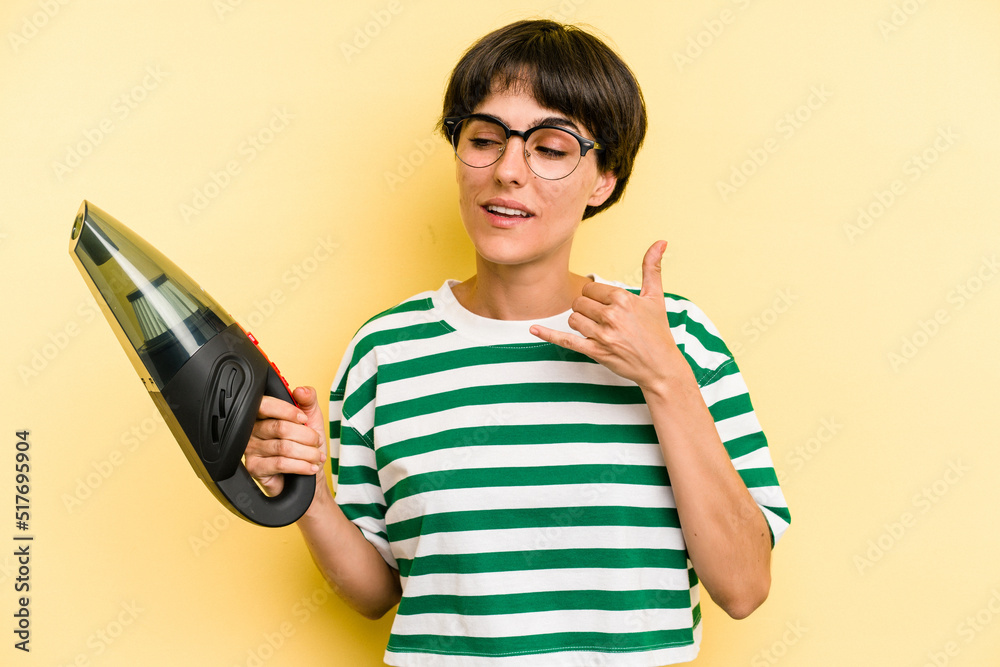 Young caucasian woman holding a hand vacuum cleaner isolated on yellow background showing a mobile phone call gesture with fingers. Young caucasian woman holding a hand vacuum cleaner isolated on yellow background showing a mobile phone call gesture with fingers.
