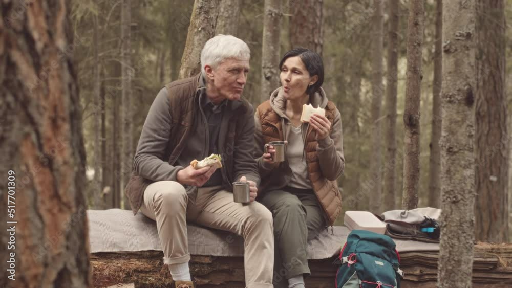 Slowmo of mature active couple with hiking backpacks sitting on log in forest, eating homemade sandwiches and drinking hot tea from thermos Slowmo of mature active couple with hiking backpacks sitting on log in forest, eating homemade sandwiches and drinking hot tea from thermos