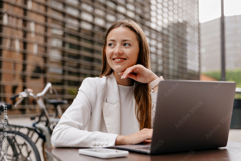 Close up portrait of smiling stylish european woman in white jacket sitting on street cafe and working on laptop. Attractive business lady in jacket rests outside