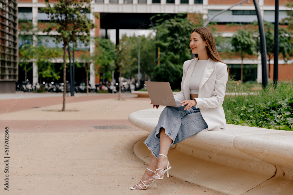 Fototapeta premium Adorable lovely girl with wonderful smile wearing white jacket and jeans sitting outdoor with laptop and working remote on background of modern building 