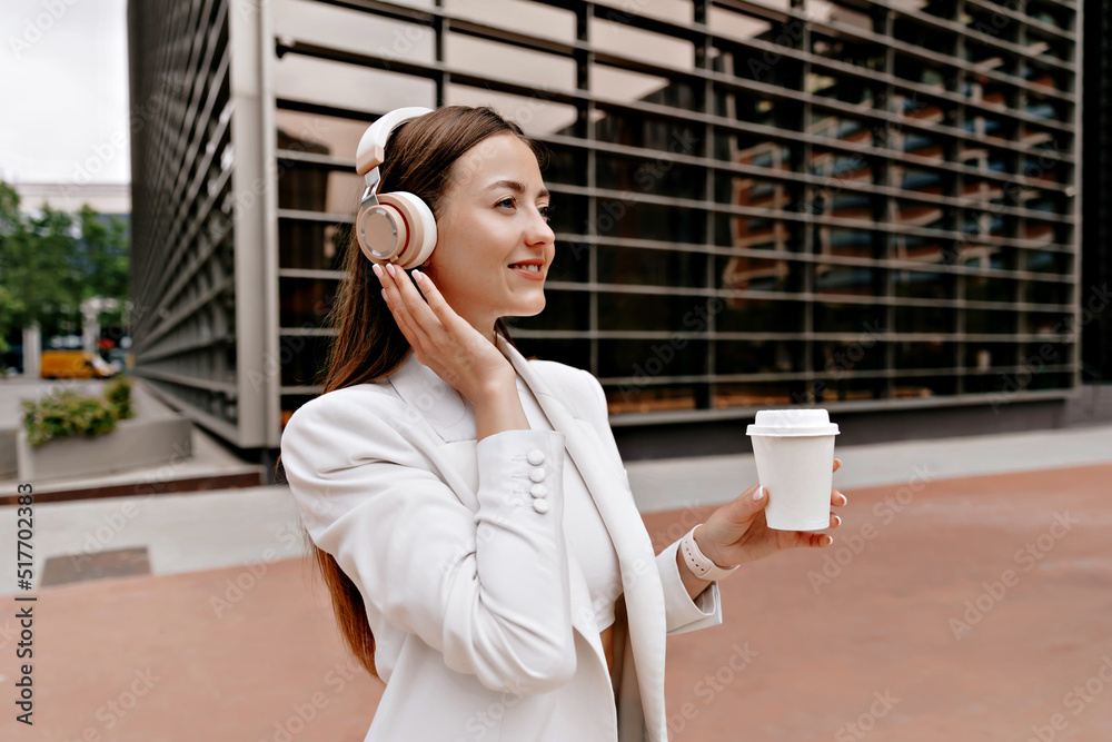 Good-looking stylish woman with long hair wearing white jacket posing with coffee in headphones on business district. Active lifestyle of business woman.